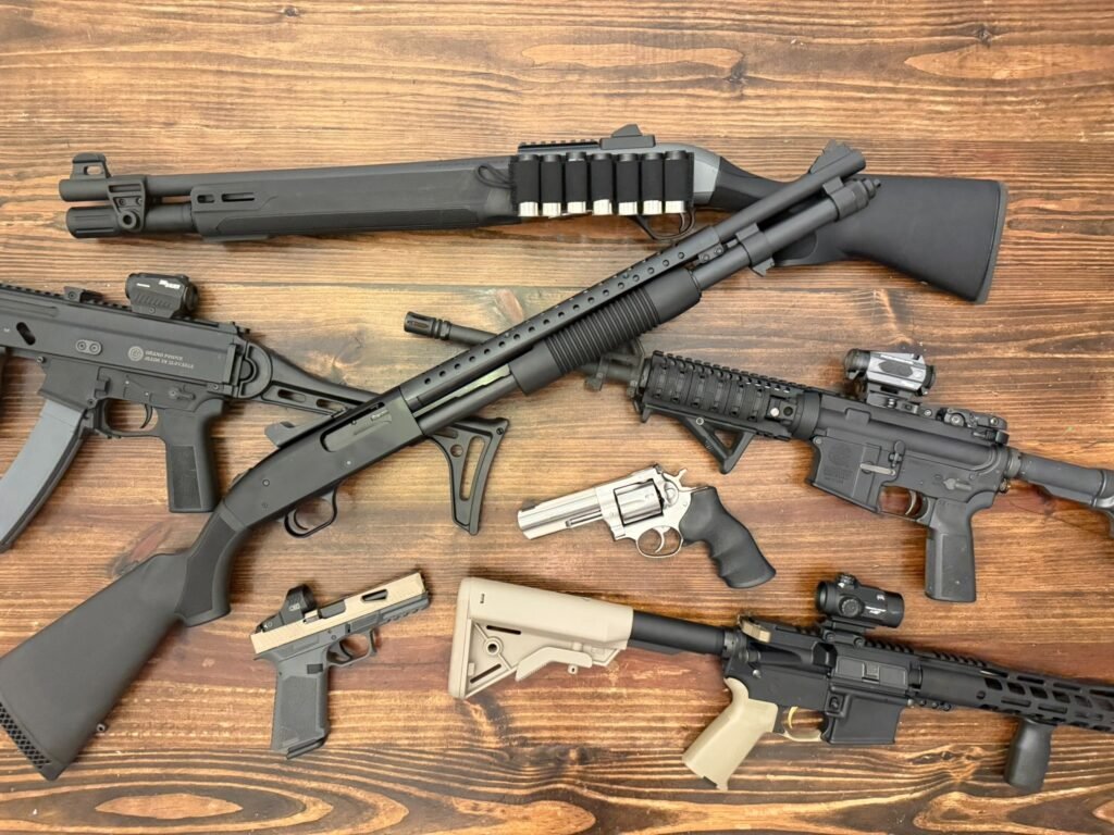 Home defense firearms including a pump shotgun, AR-15 style rifle, PCC, revolver, and compact handgun arranged on a wooden table, illustrating platform choices discussed in a home defense ammunition guide.