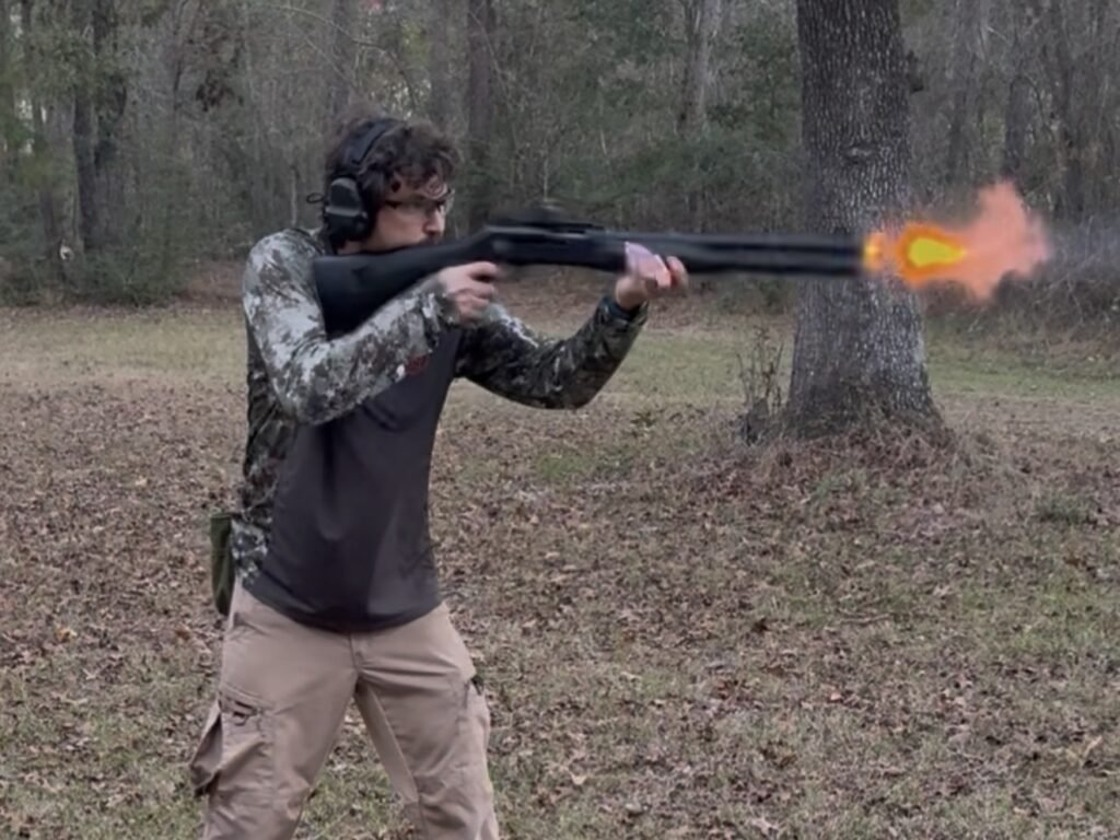 Shooter firing a semi-automatic shotgun outdoors on a wooded range, captured mid-shot with visible muzzle flash, demonstrating live-fire testing of 12-gauge buckshot.